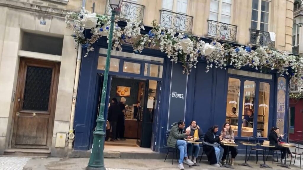 ジェフリー・カーニュの正面写真。青い外壁の前にベンチと椅子があり、ダウンを着た２人組がケーキを食べています。