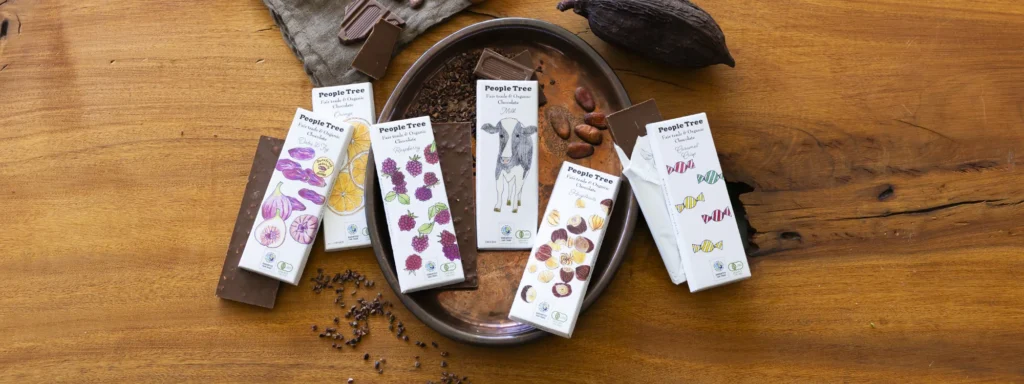 ピープルツリーの板チョコシリーズの商品画像。木製の台の上に、紙製のラッピングに包まれた全部で6個の商品が並ぶ。左からデーツフィグ、オレンジ、ラズベリー、ミルク、ヘーゼルナッツ、カラメルクリスプ。